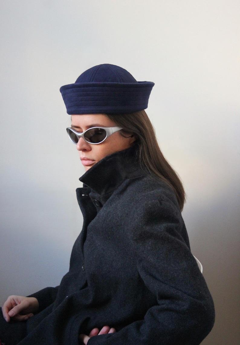 May include: A navy blue sailor hat with a structured brim. The hat is worn with a dark gray coat and silver-framed sunglasses. The hat's design is a classic style, suitable for various outfits.