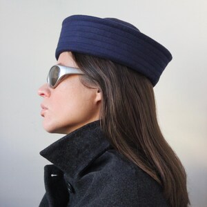 May include: A navy blue hat with horizontal ribbed detailing. The hat is worn with silver-framed sunglasses and a dark gray coat. The image is a side profile against a plain white background. The hat appears to be made of a felt-like material.