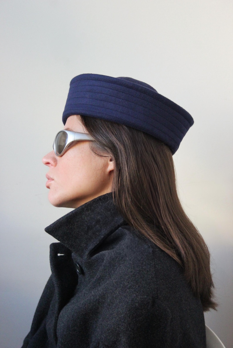 May include: A navy blue hat with horizontal ribbed detailing. The hat is worn with silver-framed sunglasses and a dark gray coat. The image is a side profile against a plain white background. The hat appears to be made of a felt-like material.