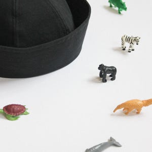 May include: A solid black sailor-style cap with a rounded crown and a wide, folded brim. The cap is surrounded by a collection of small, colorful plastic animal figurines, including a zebra, gorilla, turtle, and various other wildlife species.