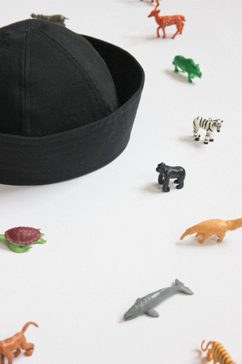 May include: A solid black sailor-style cap with a rounded crown and a wide, folded brim. The cap is surrounded by a collection of small, colorful plastic animal figurines, including a zebra, gorilla, turtle, and various other wildlife species.