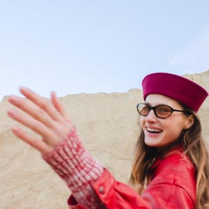 May include: A woman wearing a red jacket, a burgundy hat, and black-framed glasses smiles. Her sleeve shows a red and white patterned cuff. The background features a sandy landscape and a clear sky.