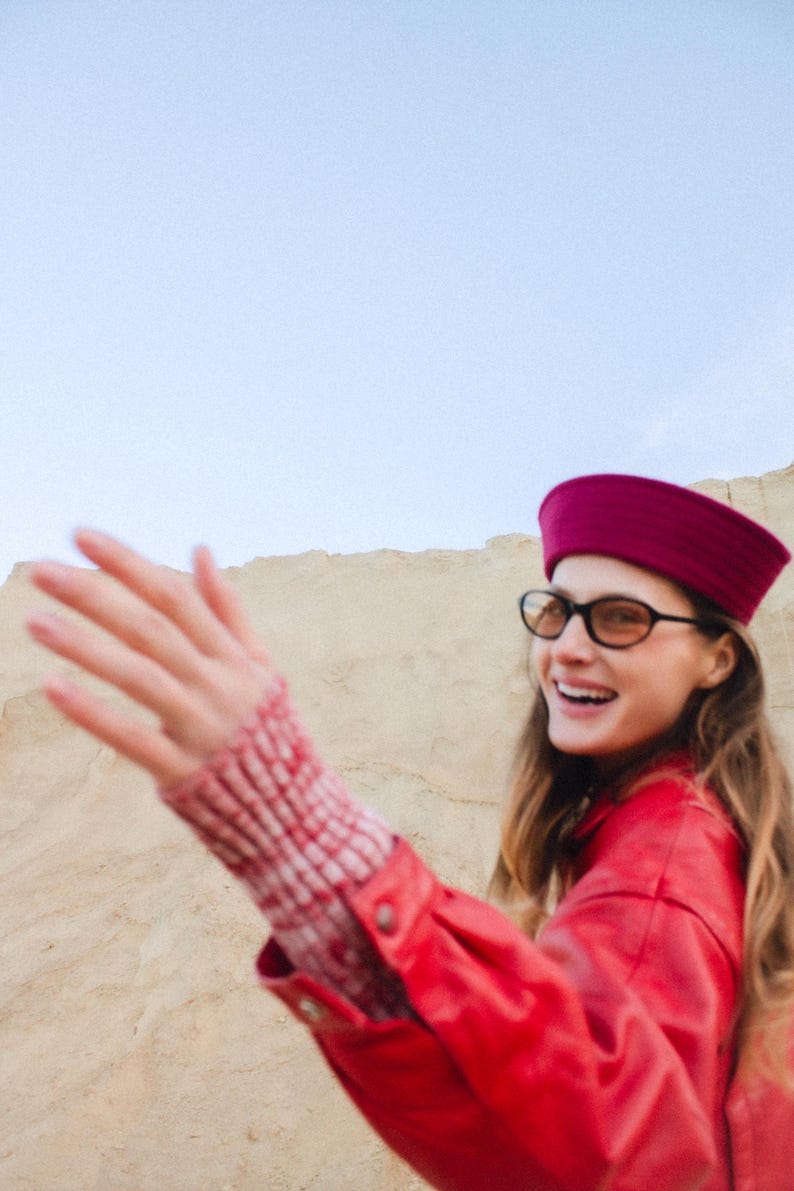 May include: A woman wearing a red jacket, a burgundy hat, and black-framed glasses smiles. Her sleeve shows a red and white patterned cuff. The background features a sandy landscape and a clear sky.