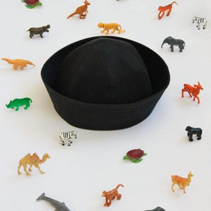 May include: A solid black sailor-style cap with a rounded crown and a wide, turned-up brim, surrounded by a collection of small, colorful plastic animal figurines including a zebra, elephant, camel, monkey, tiger, and various sea creatures.