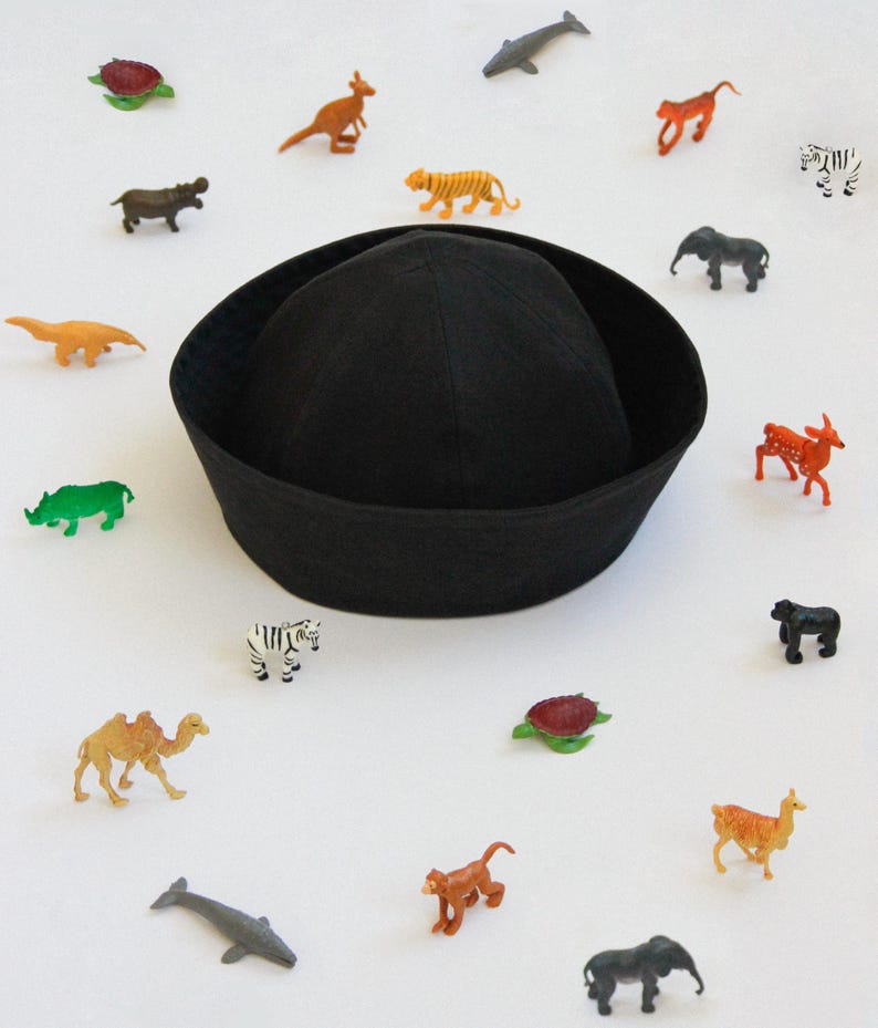 May include: A solid black sailor-style cap with a rounded crown and a wide, turned-up brim, surrounded by a collection of small, colorful plastic animal figurines including a zebra, elephant, camel, monkey, tiger, and various sea creatures.