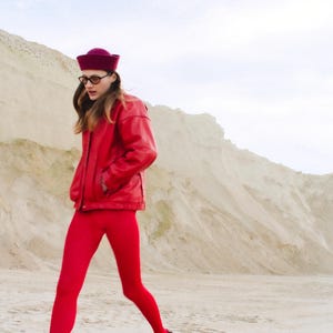 May include: A person wearing a red leather jacket, matching leggings, and a burgundy hat. The outfit is paired with black shoes and glasses. The background features a sandy landscape and a cloudy sky.