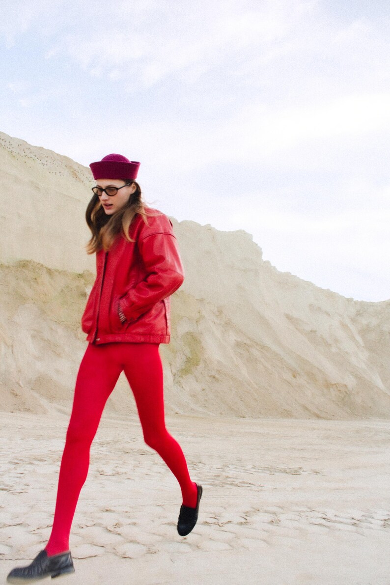 May include: A person wearing a red leather jacket, matching leggings, and a burgundy hat. The outfit is paired with black shoes and glasses. The background features a sandy landscape and a cloudy sky.