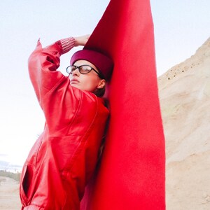 May include: A person wearing a red leather jacket, matching leggings, and a burgundy hat and glasses, leaning against a large, bright red fabric. The background features a desert landscape and a clear sky.