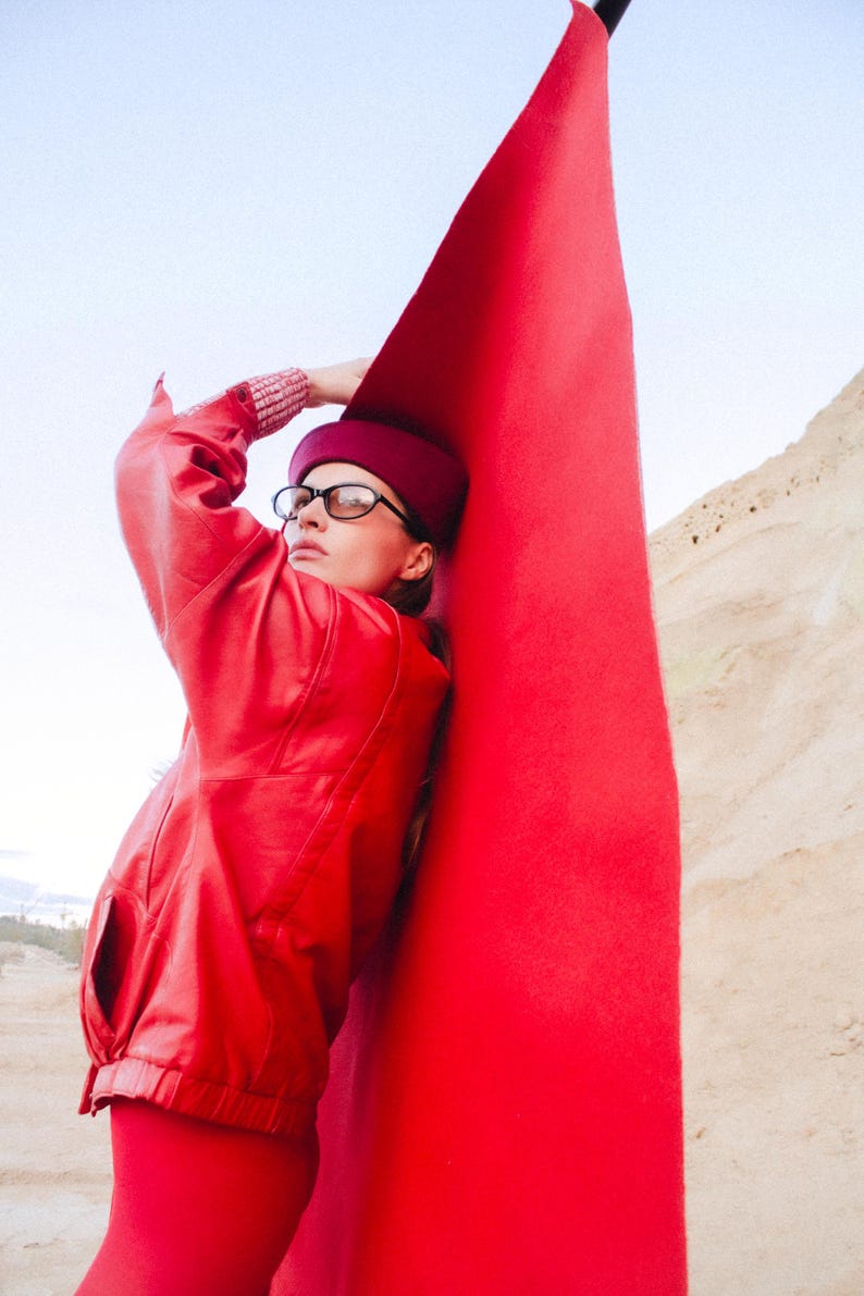 May include: A person wearing a red leather jacket, matching leggings, and a burgundy hat and glasses, leaning against a large, bright red fabric. The background features a desert landscape and a clear sky.