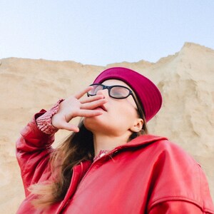 May include: A person wearing a red leather jacket, a burgundy hat, and black-framed glasses. The person is shielding their eyes with their hand, with a sandy background.