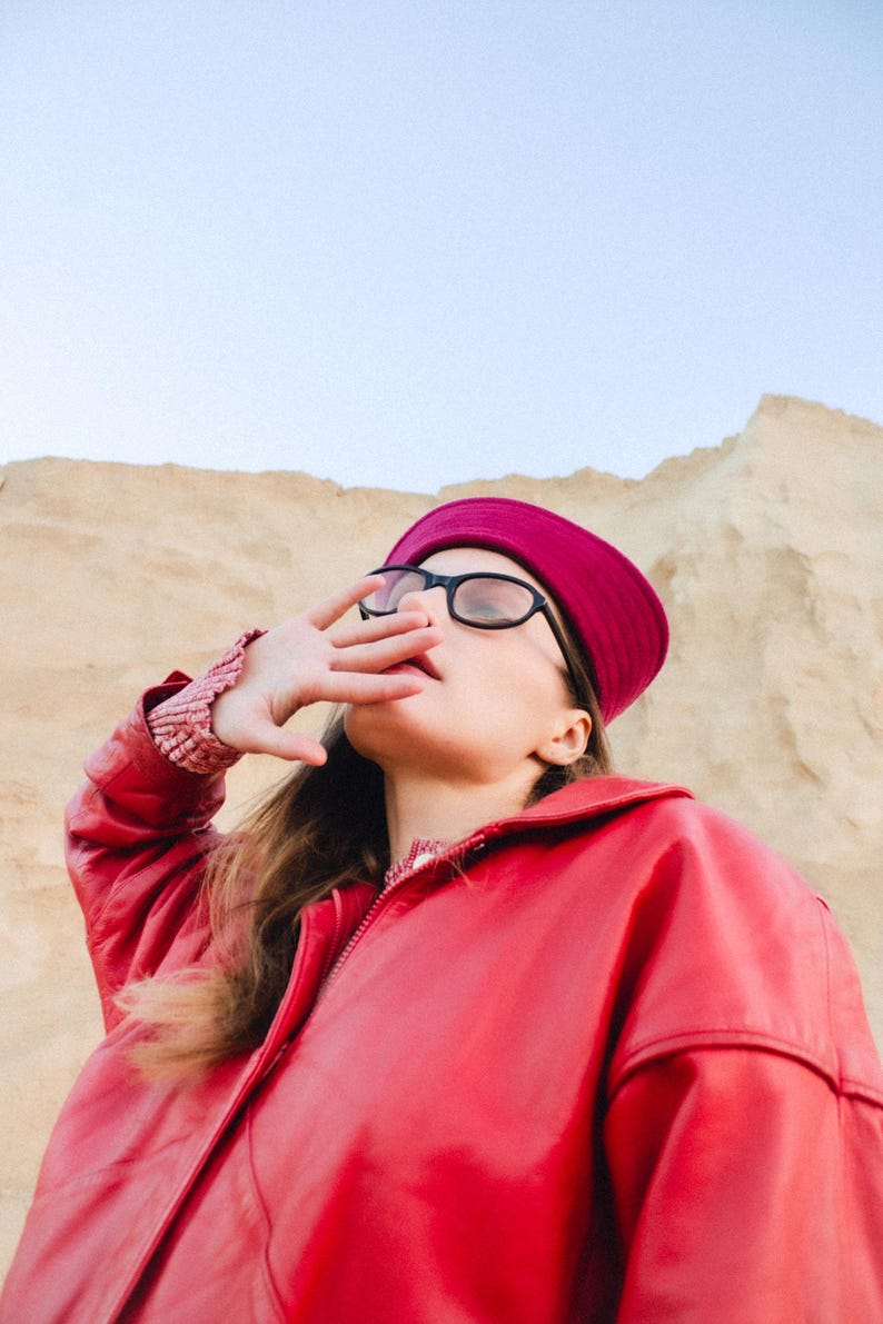 May include: A person wearing a red leather jacket, a burgundy hat, and black-framed glasses. The person is shielding their eyes with their hand, with a sandy background.
