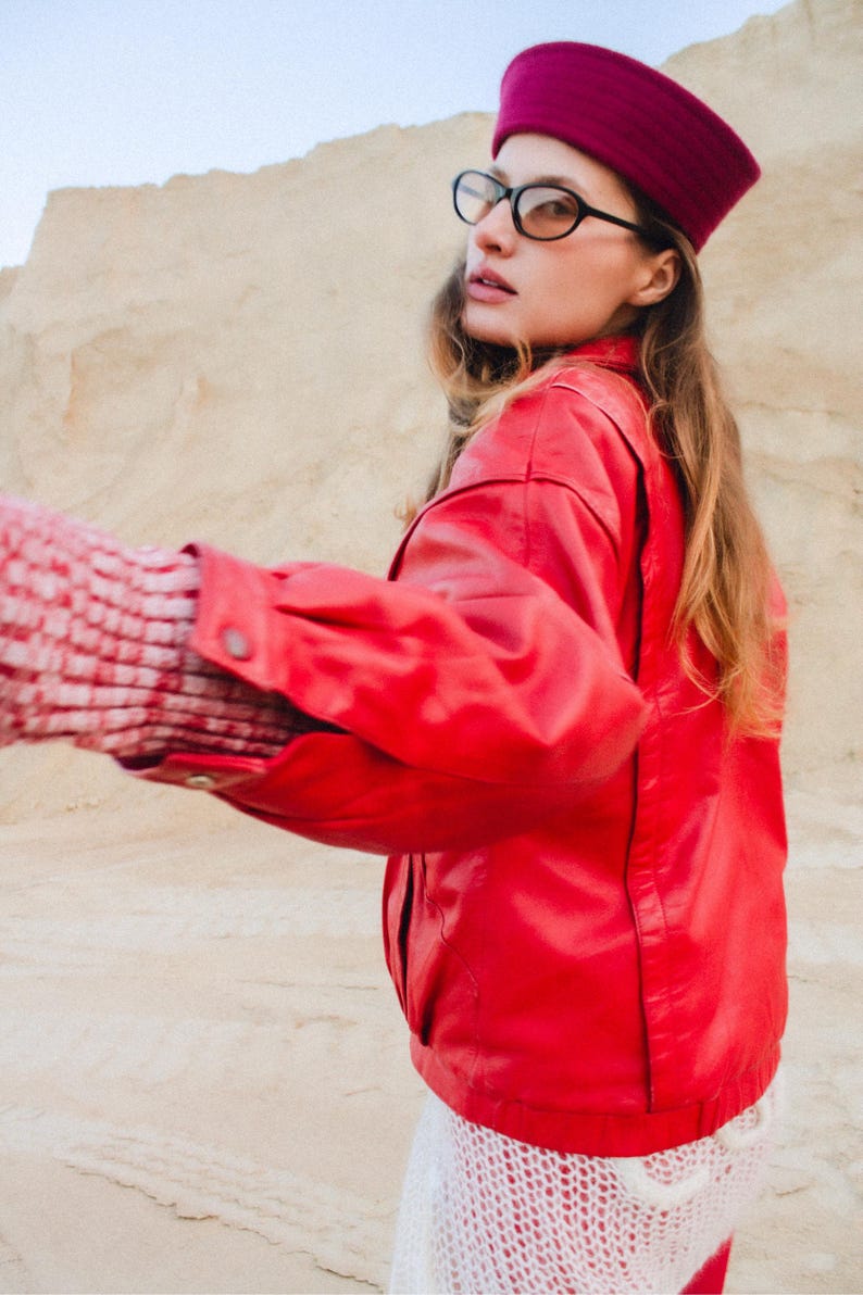 May include: A bright red jacket with a matching maroon hat and black-framed glasses. The jacket has a classic bomber style. The sleeves are cuffed, and the jacket is paired with a white, open-weave garment.