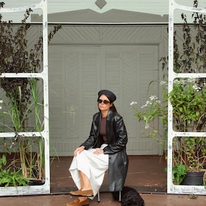 May include: A black beret, dark sunglasses, a brown top, a long black leather coat, a white skirt, and brown suede loafers. A small black dog is resting on the ground nearby. The outfit is worn by a person sitting on a chair.