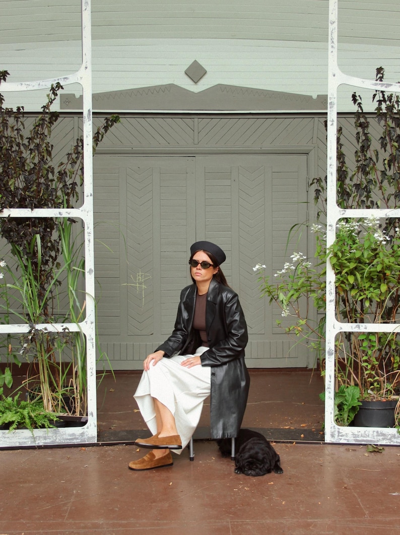 May include: A black beret, dark sunglasses, a brown top, a long black leather coat, a white skirt, and brown suede loafers. A small black dog is resting on the ground nearby. The outfit is worn by a person sitting on a chair.