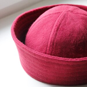 May include: A close-up shot of a deep red hat with a rounded crown and a brim. The hat is made of a soft, felt-like material. The hat is sitting on a white surface.