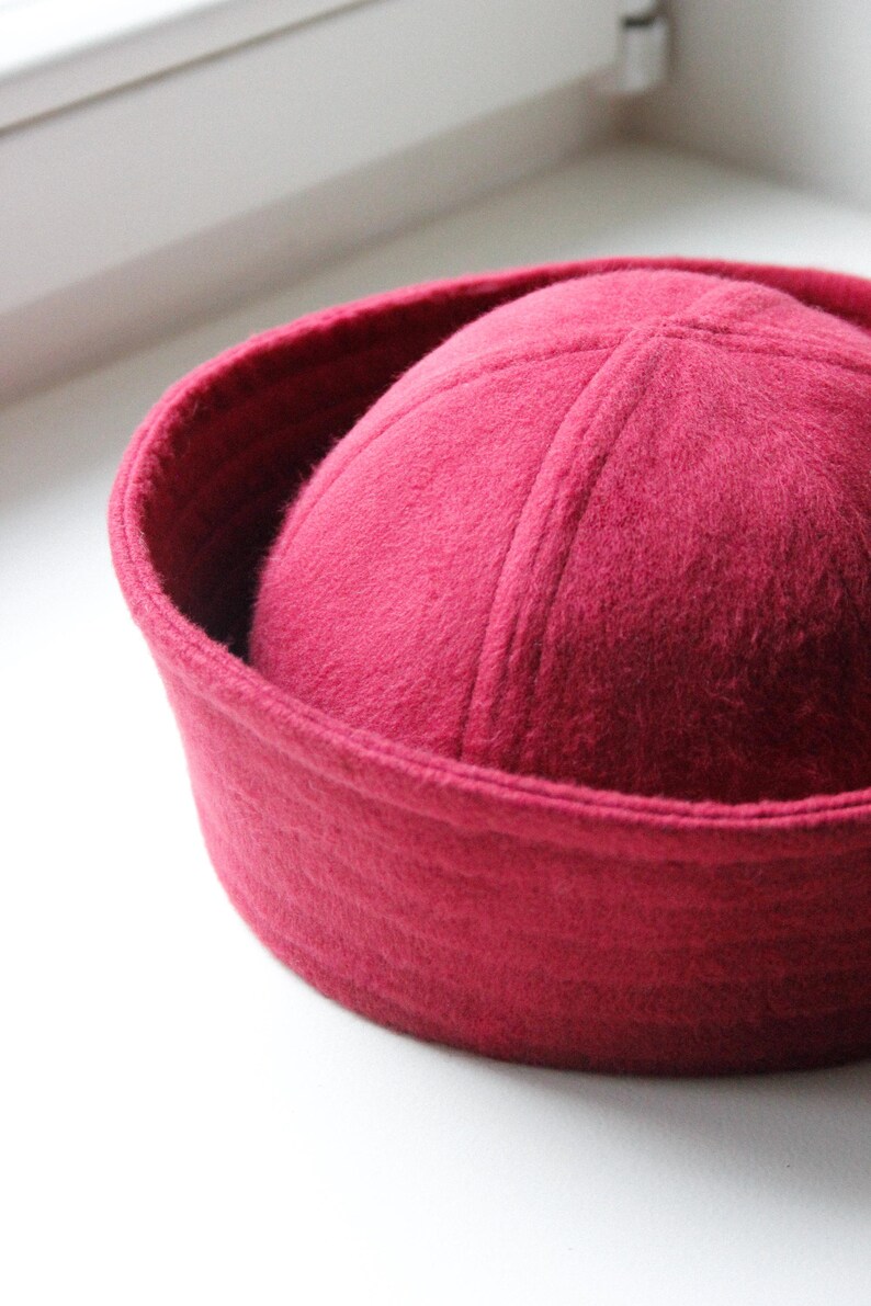 May include: A close-up shot of a deep red hat with a rounded crown and a brim. The hat is made of a soft, felt-like material. The hat is sitting on a white surface.
