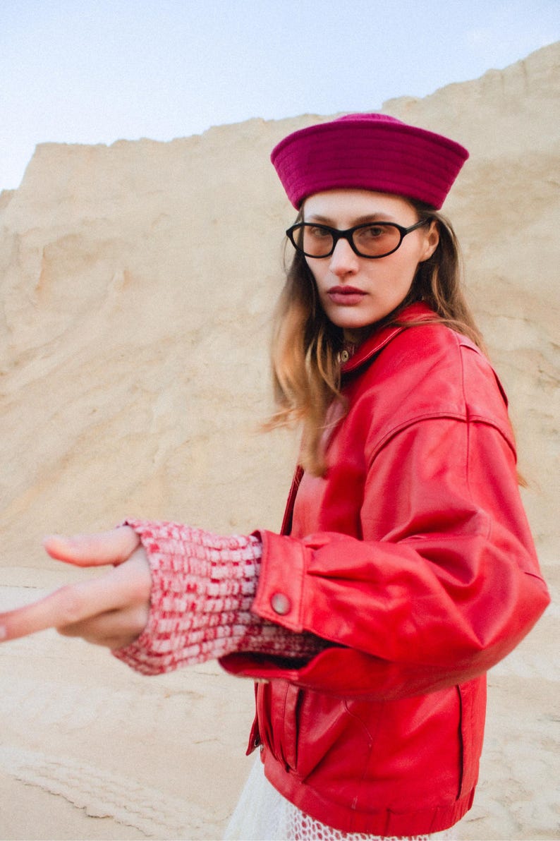 May include: A woman wearing a red leather jacket, a fuchsia hat, and black-framed glasses. The jacket has a buttoned cuff and a collared neckline. The sleeves of the jacket are over a red and white patterned sleeve. The background is a sandy landscape.