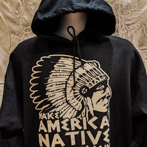 May include: Black hooded sweatshirt with a large graphic of a Native American chief in profile, wearing a headdress. The text "MAKE AMERICA NATIVE AGAIN" is printed below the image in a bold, sans-serif font. The sweatshirt has a front pocket and drawstring hood.
