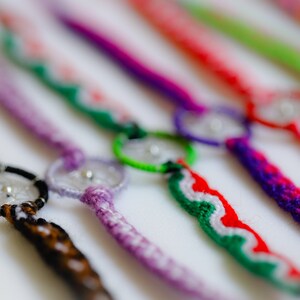 May include: Close-up of several handmade friendship bracelets in various colors. Each bracelet features a central dreamcatcher design with a small silver bead. The bracelets are made with woven threads in shades of brown, purple, red, and green.
