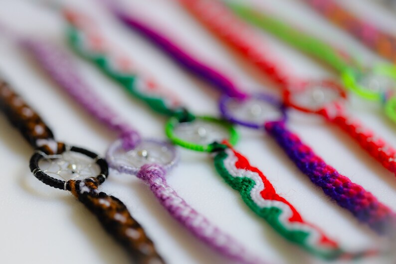 May include: Close-up of several handmade friendship bracelets in various colors. Each bracelet features a central dreamcatcher design with a small silver bead. The bracelets are made with woven threads in shades of brown, purple, red, and green.