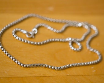 Op de afbeelding: Een zilveren ketting met een box chain-ontwerp. De ketting ligt op een houten oppervlak.