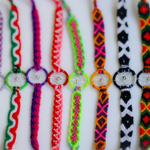 May include: A collection of colorful friendship bracelets with dreamcatcher charms. The bracelets feature various woven patterns in shades of brown, purple, red, green, orange, pink, black, and white. Each bracelet has a small, silver-toned dreamcatcher.
