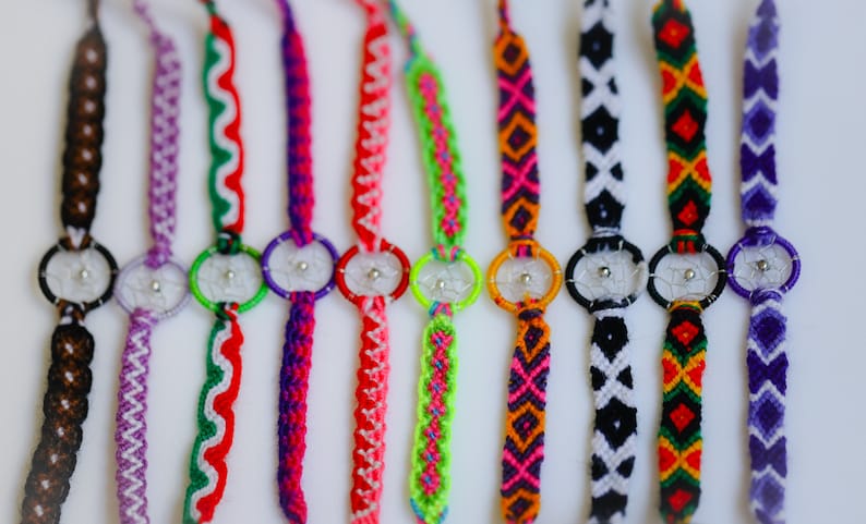 May include: A collection of colorful friendship bracelets with dreamcatcher charms. The bracelets feature various woven patterns in shades of brown, purple, red, green, orange, pink, black, and white. Each bracelet has a small, silver-toned dreamcatcher.
