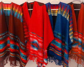 May include: Four colorful ponchos with geometric patterns and fringe. The ponchos are displayed on hangers and are in shades of brown, red, blue, and orange.