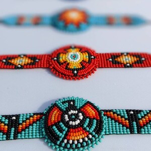 May include: Four beaded bracelets with colorful geometric patterns. The bracelets are made with small, round beads in shades of red, orange, yellow, blue, black, and white. The bracelets are arranged in a row, with the top bracelet out of focus and the bottom bracelet in focus.