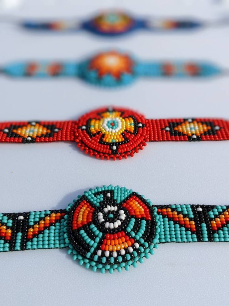 May include: Four beaded bracelets with colorful geometric patterns. The bracelets are made with small, round beads in shades of red, orange, yellow, blue, black, and white. The bracelets are arranged in a row, with the top bracelet out of focus and the bottom bracelet in focus.