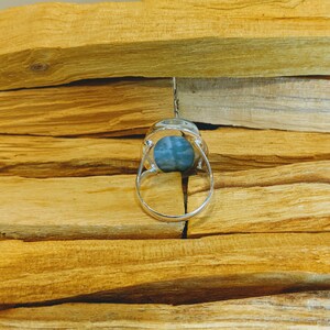 May include: A silver ring with a blue-green gemstone set in a bezel setting. The ring is on a wooden surface.