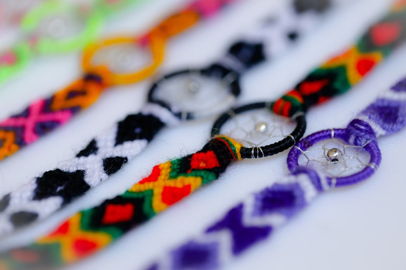 May include: Close-up of several handmade friendship bracelets with dreamcatcher charms. The bracelets feature woven patterns in various colors, including orange, black, purple, and green. Each bracelet has a small, silver-toned dreamcatcher charm.