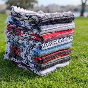 Puede incluir: Una pila de mantas tejidas de colores con rayas negras, blancas, rojas y azules. Las mantas están dobladas y apiladas unas sobre otras en una superficie de hierba verde.