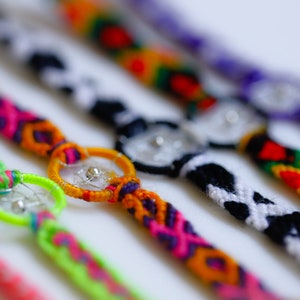 May include: Close-up of several colorful handmade friendship bracelets. Each bracelet features a unique design with vibrant colors like pink, green, orange, and purple. Some bracelets have a small, decorative charm in the center.