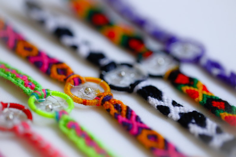 May include: Close-up of several colorful handmade friendship bracelets. Each bracelet features a unique design with vibrant colors like pink, green, orange, and purple. Some bracelets have a small, decorative charm in the center.