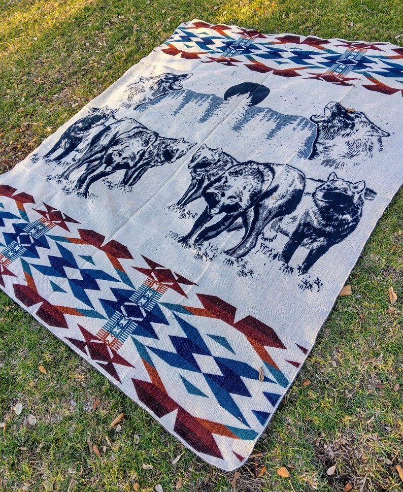 May include: A white woven blanket with a blue, red, and brown geometric pattern. The blanket features a scene of wolves howling at the moon in a forest.