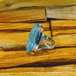 May include: A silver ring featuring a light blue oval-shaped gemstone. The ring is displayed on a wooden surface.