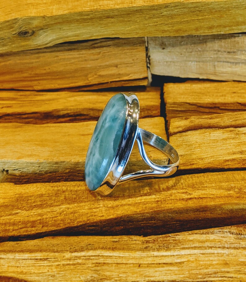 May include: A silver ring featuring a light blue oval-shaped gemstone. The ring is displayed on a wooden surface.