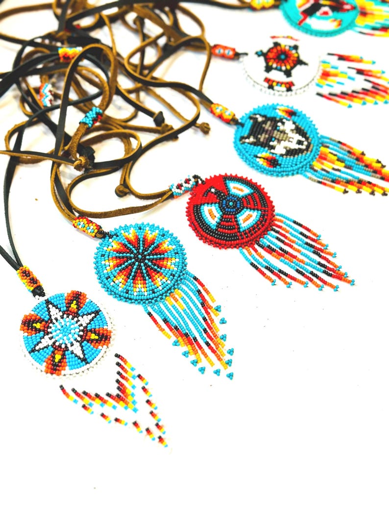 May include: A collection of beaded necklaces with colourful, geometric designs. Each necklace features a round pendant with a different pattern, including a star, a wolf, and a circle. The pendants are attached to leather cords.
