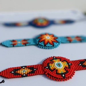 May include: Three beaded bracelets with colorful geometric patterns. The bracelets are red, blue, and turquoise with black and white accents. The center of each bracelet features a circular design with a starburst pattern.