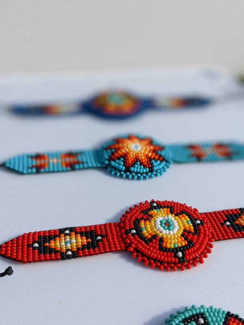 May include: Three beaded bracelets with colorful geometric patterns. The bracelets are red, blue, and turquoise with black and white accents. The center of each bracelet features a circular design with a starburst pattern.