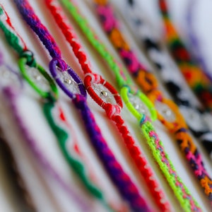 May include: A collection of colorful friendship bracelets. Each bracelet features a woven design in various colors, including red, purple, green, and orange. The bracelets have a small, clear bead in the center.