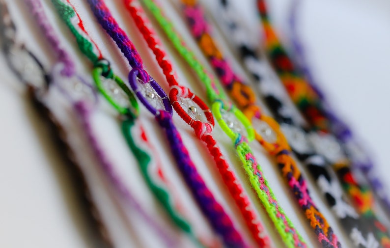 May include: A collection of colorful friendship bracelets. Each bracelet features a woven design in various colors, including red, purple, green, and orange. The bracelets have a small, clear bead in the center.