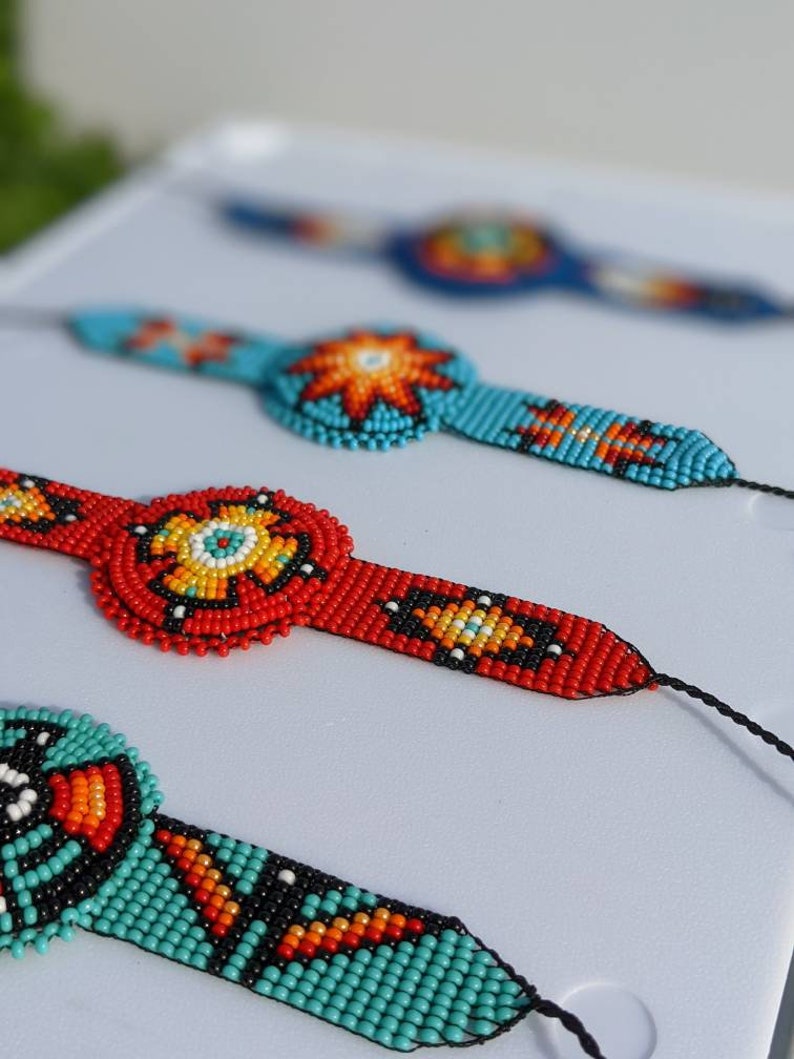 May include: Three beaded bracelets with colorful geometric patterns. The bracelets are made with small, colorful beads and feature a central circular design. The bracelets are displayed on a white surface.
