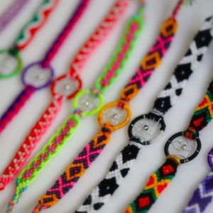 May include: A collection of colorful friendship bracelets with woven patterns and dreamcatcher charms. The bracelets feature various colors including red, pink, green, black, and purple. Each bracelet has a small, silver-colored dreamcatcher charm in the center.