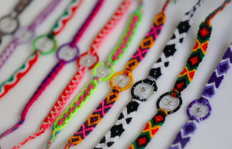 May include: A collection of colorful friendship bracelets with woven patterns and dreamcatcher charms. The bracelets feature various colors including red, pink, green, black, and purple. Each bracelet has a small, silver-colored dreamcatcher charm in the center.