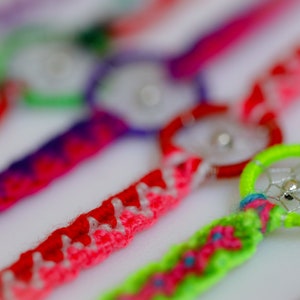 May include: Close-up of colorful dreamcatcher bracelets. The bracelets feature woven bands in various colors, including red, pink, purple, and green. Each bracelet has a small dreamcatcher charm with a silver bead in the center.