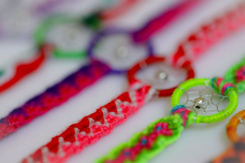 May include: Close-up of colorful dreamcatcher bracelets. The bracelets feature woven bands in various colors, including red, pink, purple, and green. Each bracelet has a small dreamcatcher charm with a silver bead in the center.