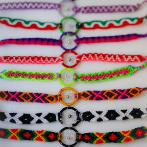 May include: A collection of colorful handmade friendship bracelets. Each bracelet features a woven design in various colors, including red, green, purple, and black, and incorporates a small dreamcatcher charm. The bracelets are displayed on a white surface.