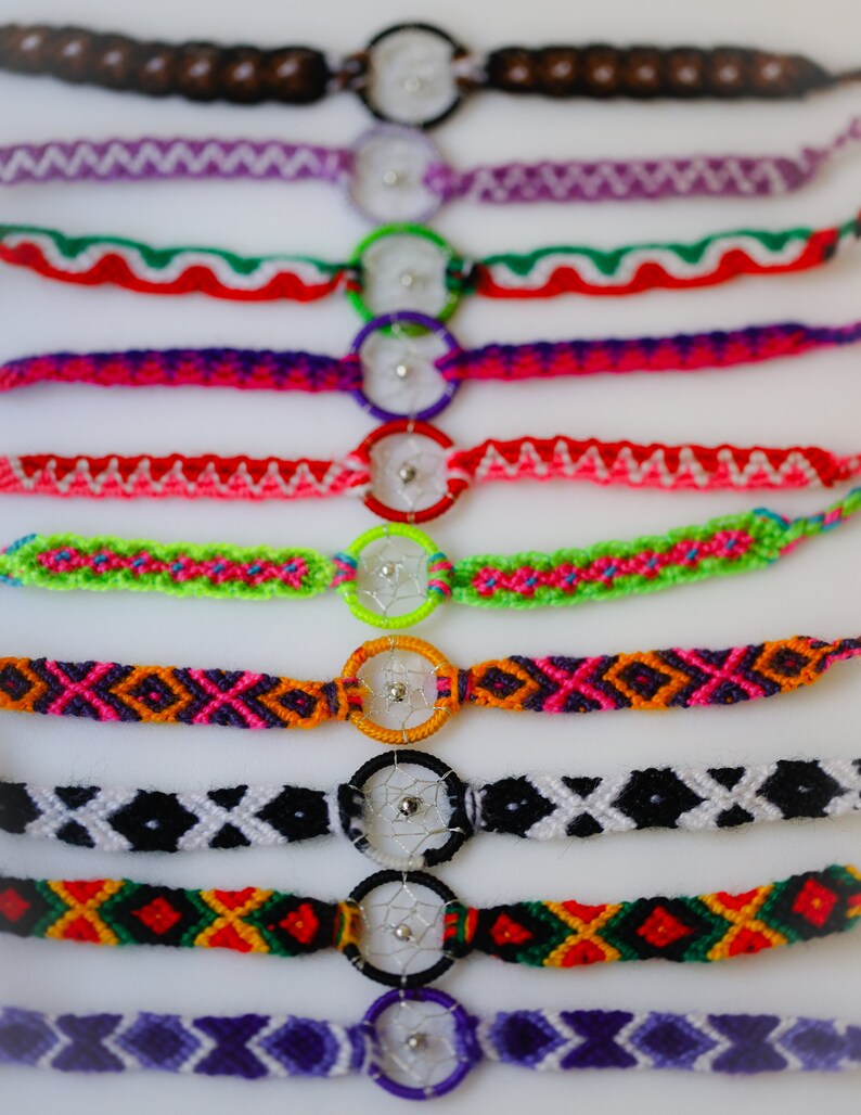 May include: A collection of colorful handmade friendship bracelets. Each bracelet features a woven design in various colors, including red, green, purple, and black, and incorporates a small dreamcatcher charm. The bracelets are displayed on a white surface.